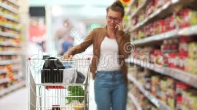 一个年轻的现代女孩正在超市购物，用手机聊天。 真诚的微笑。 超市手推车是